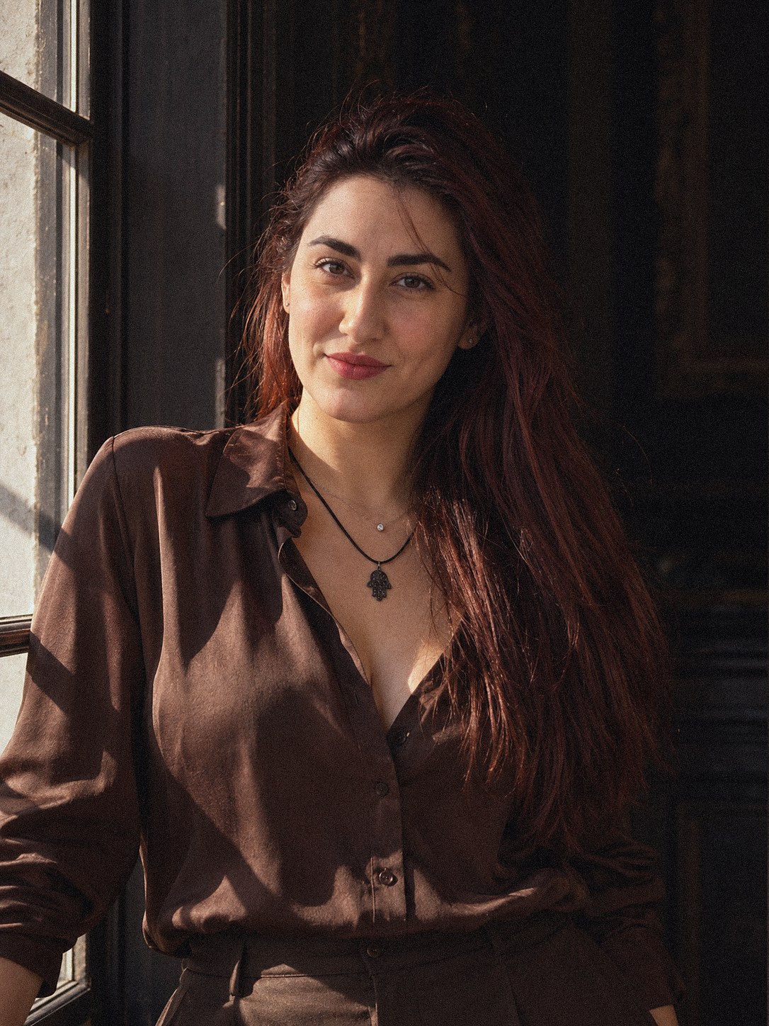 Retrato de Rachel Cossich, de camisa marrom, sorrindo ao lado de uma janela com luz do sol